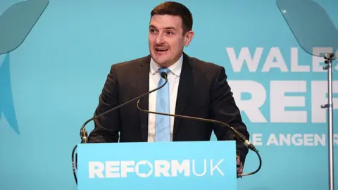 James Evans at lectern with Reform UK banner across the front at a rally speaking into microphone wearing black suit and white shirt and light blue tie. He has short dark hair.