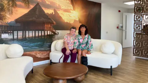 Jon Wright/BBC Two women sit on a sofa. There is a big piece of art on the wall of a sunny tropical scene.