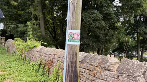 A poster reading SUPPORT OUR OBJECTON OFFERTON LANE DOES NOT WANT PADEL is attached to wooden lamp post near a stone fence in Offerton.