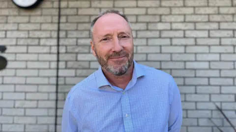 BBC Man in his 50s outside the Les Varandes site. He has a grey beared and grey hair which is balding at the front. 
