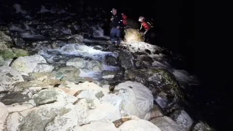 WMRT Mountain Rescue volunteers, wearing head torches and emergency gear, walk over a swollen waterway which crosses a path. The two members were deployed in the search for a young walker. 