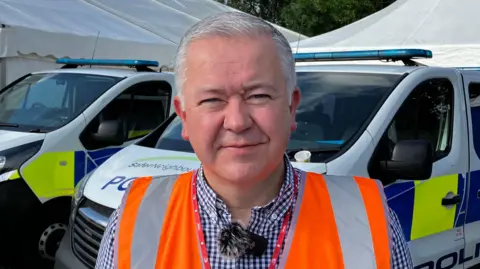 Matt Marvel/BBC Det Supt Matt Bodmer smiles at the camera. He has short grey hair and wears a chequered shirt with an orange hi-vis jacket over the top. Police vans are parked up behind him at an event. 