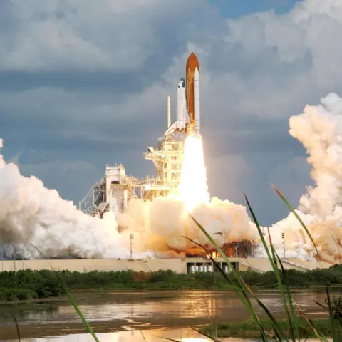 NASA Space Shuttle during launch at Cape Canaveral in Florida. The white shuttle is vertically attached to an orange rocket with large white booster engines. Flames emit from the rocket engine and a large amount of white smoke appears as the rocket is about to take off. There is a lake in the foreground.