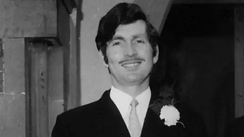 Doherty Family A black-and-white photo of a man wearing a suit with a white flower corsage pinned to his lapel. He has a thin moustache and dark, side-parted hair.