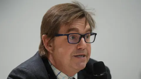 Nik Johnson (left), Mayor of Cambridgeshire and Peterborough, wearing a checked shirt and blue suit speaking into a microphone.