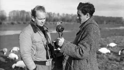 Peter Scott, the Director of the Severn Wild Fowl Trust, photographed at the headquarters of the Trust, Slimbridge, with Desmond Hawkins, BBC Features producer, West Region. Mr. Hawkins is holding a microphone which has "The BBC" written on it