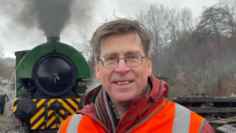 A man wearing a hi-vis jacket stands in front of a green and black steam train.
