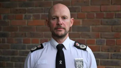 Jason Hogg is wearing his Thames Valley Police shirt and black tie, and is stood in front of a brick wall.