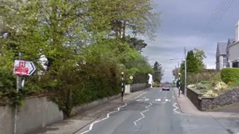 The road junction at Ramsey Cottage Hospital. There is a road sign with an H on it pointing to the hospital to the left and a pelican crossing up ahead across the road.