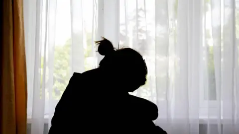 A silhouette of a woman in front of a window, which has a curtain drawn across it.