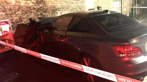 A grey BMW car has collided with an old stone wall. There is red and white cordon tape in the foreground.