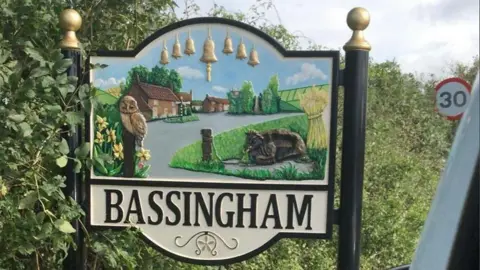 A large village sign on the road into Bassingham. An image depicts houses, a road, a green, a cow, an owl, trees, flowers and symbols of village life, including a sheaf of wheat and church bells. A 30mph road sign can be seen in the background.