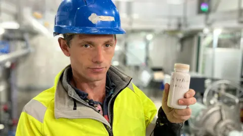 A man in high-viz and hard hat, holds a small bottle containing a high density liquid that is being used as part of a pilot pumped hydro power project near Plymouth, Devon
