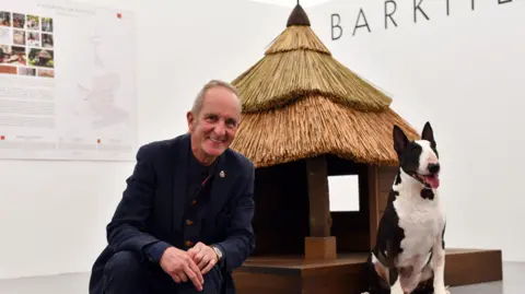 PA Media A balding man in a blue suit sits beside a thatched kennel, alongside a bull terrier.