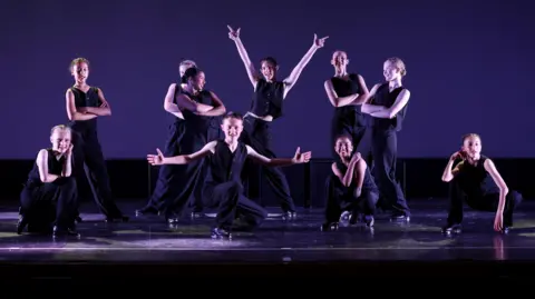 David Felton Photography A group of dancers, all dressed in black, taking part in a dance number. Six are standing and four are crouched. One girl in the middle has her arms up. They are all striking a pose. 