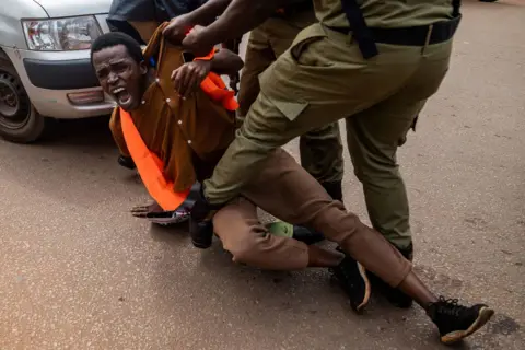 BADRU KATUMBA / AFP Ugandan police arrest an environmental activist during a protest in Kampala against the East African Crude Oil Pipeline Project. The protester lies on the ground with his mouth open as if he were screaming while police officers hold him by his shirt.