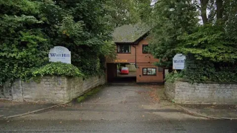 The entrance to a care home, with stone walls either side. Signs either side of the road say Wall Hill Care Home in blue lettering on a white background.