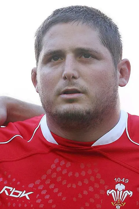 Getty Images Rhys wearing his Welsh rugby jersey singing the national anthem. He has a serious look. 
