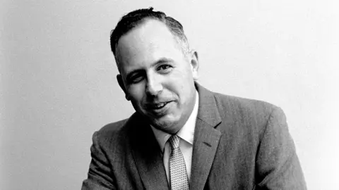 Getty Images A black-and-white mid-shot of a 39-year-old man with short, greying dark hair, dressed in a suit with a grey jacket, light-coloured shirt, and a tie with a dotted pattern on it. He is looking into the camera, smiling, leaning forward slightly, posed against a plain, featureless background.