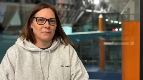 A woman is sitting down and looking straight at the camera - you can only see her upper body. She has shoulder-length brown hair, black rimmed glasses and is not smiling. She is wearing a light grey hooded top and behind her is the slightly blurred image of the inside of a leisure centre.