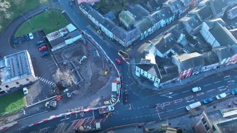 An aerial view of roadworks in Workington town. There are red and white striped barriers around a large construction area on the left of the image and traffic cones separating the road lanes.