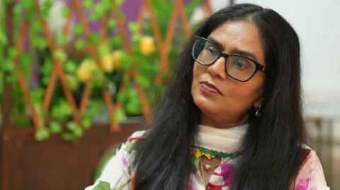 A South Asian lady with long, straight black hair and black thick rimmed glasses. She wears a white traditional outfit with colourful patterning