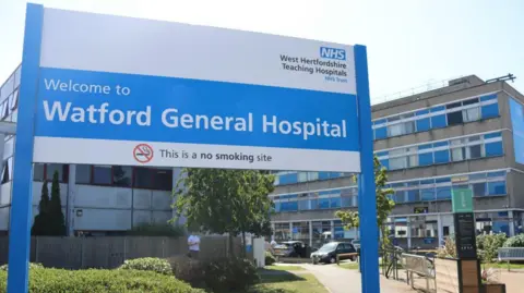 A large blue and white sign is in the foreground saying Welcome to Watford General Hospital" with the NHS logo above it. The hopsital - a concrete block of 4 storeys -is behind.