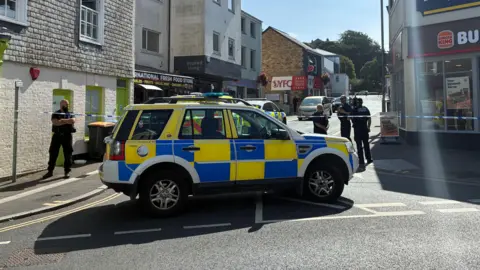 BBC A police Range Rover and several officers are stationed at the end of a cordoned off road in Truro, Cornwall. Blue and white police tape has been spread across the street. A Burger King restaurant is at the edge of the cordon.