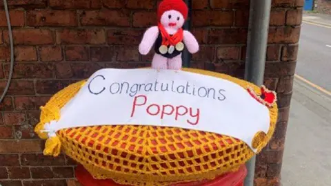 A postbox topper showing a knitted doll wearing a swimming cap and three medals around its neck. The cover is gold and has the words "Congratulations Poppy".  