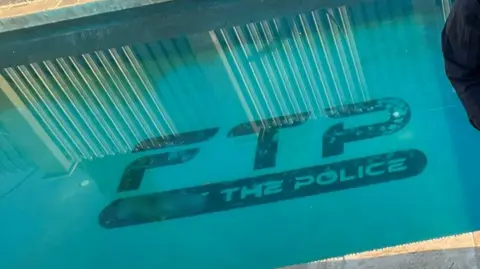 ERSOU A swimming pool with clear blue water. At the bottom of the pool, large black letters spell out “FTP” with smaller text underneath reading “THE POLICE.” Vertical metal bars and part of a poolside area are visible at the top edge.