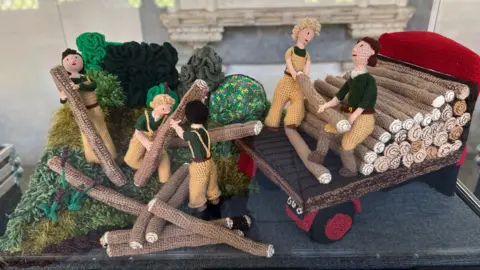 Knitted figures are carrying logs onto a wagon.