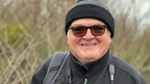 Pictured is Kevin Clements, a man with dark glasses, a black woolly hat, a black coat and a camera strap around his neck. He is smiling and looking right at the camera.