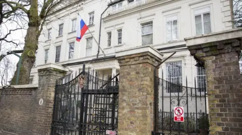 External image of the Russian embassy building in London - a white building behind a stone fence with large black wired gate.