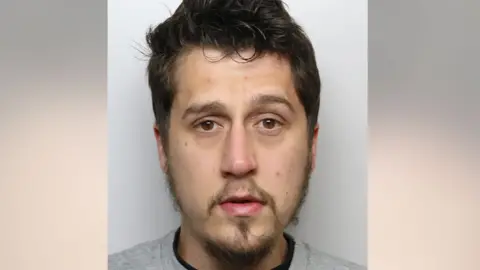 A man with dark hair and a goatee beard wearing a police issue grey sweatshirt 