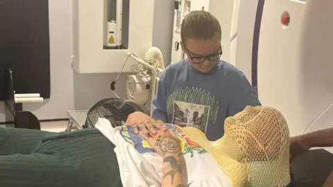 Libby Woolaston The image shows a person lying on a hospital treatment table inside a clinical radiotherapy or scanning room. The person is wearing a rigid mesh head mask and is positioned carefully on the bed, suggesting preparation for head‑focused treatment. Medical equipment and control panels surround the table. Another person stands beside the bed, looking down at the patient, with one hand resting near the patient’s arm.