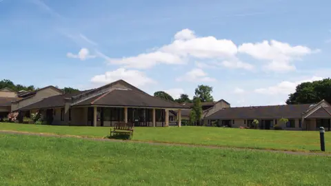 The picture shows a wide outdoor view of a low, spread‑out brown building complex set in a large grassy area. The buildings appear to be single storey, with pitched roofs covered in dark tiles and light‑coloured walls. 