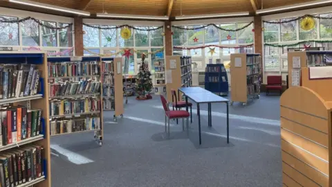 Laura Coffey/BBC Interior of single-storey library building with windows all round the edge of the hexagonal building. There are wooden shelves full of books, a study table and a counter to the right. There are Christmas decorations on display.