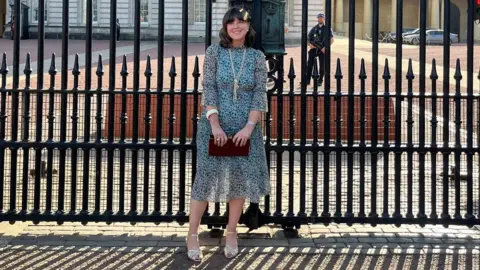 Caroline Jones Caroline Jones stands outside the gates of Buckingham Palace. She wears a green floral dress and gold sandals. She is carrying a brown clutch bag. She has brown shoulder length hair with a fringe. An armed guard stands behind her in the palace grounds. He is looking on.