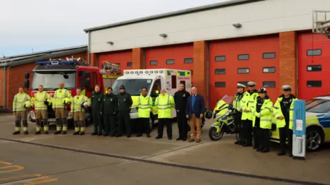 Suffolk Police Paramedics, police officers, and firefighters standing in a row outside a fire station and in front of a police car, a fire engine and an ambulance. 