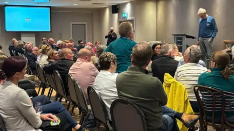 People sit in a conference room. A man in a blue jumper is on a stage and a woman in green is standing in the audience and speaking to him.