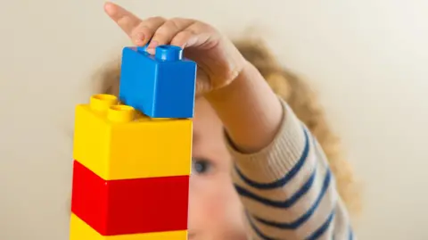 A close-up of a child stacking plastic toy bricks on top of one another. They wear a white and blue striped jumped and have curly blond hair