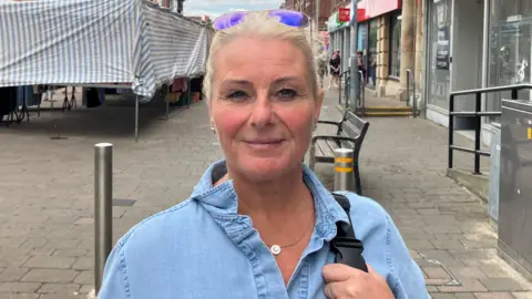 Julie Harriman, who has blonde hair which has been tied up and brown eyes. She is wearing a denim shirt and smiling in the photo. Behind her an empty shop unit can be seen.