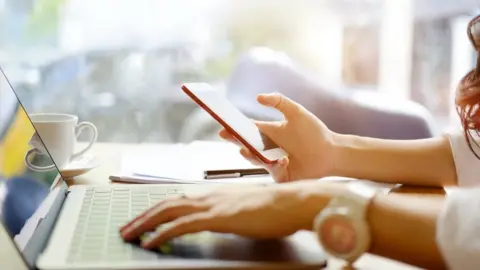 Getty Images Young woman looking at smartphone while using laptop