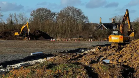 A building site with flat grey hardcore surface and mud piles has two yellow excavators sat either side of it.