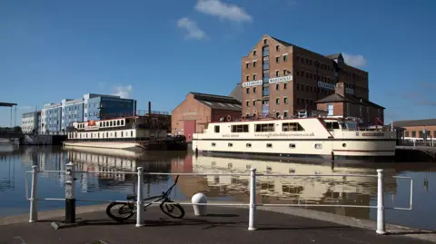 Gloucester Docks on a sunny day, with two canal cruise boats passing down the water in front of an old warehouse building and a more modern college building