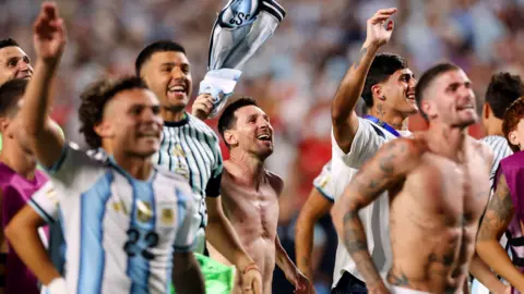 Reuters The Argentina national football team celebrating after their win against Switzerland. Central figure Lionel Messi is bare chested and waving his football shirt above his head. Other players appear to be jumping up and down, their arms aloft. All are smiling.