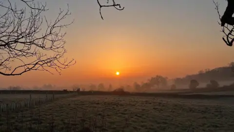 Andy Howard/BBC A sunrise with frosted fields in the foreground in the Cotswolds. Lines of hedgerows and trees are dimly visible in the haze and the sun is bright yellow with an orange haze around it