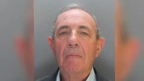 A mugshot of John Allen. He wearing dark jacket and shirt and standing against a grey background. 