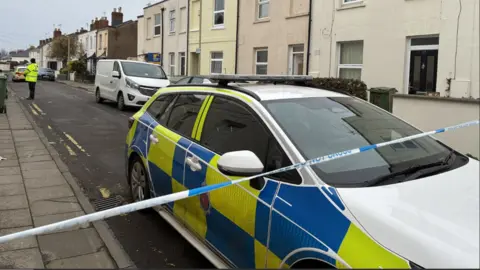 A police cordon on a residential street with a police car behind blue and white evidence tape and an officer standing guard halfway down the road