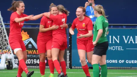 BBC Jersey player dressed in red hug whilst a Guernsey player in green looks on. They are near the goal.